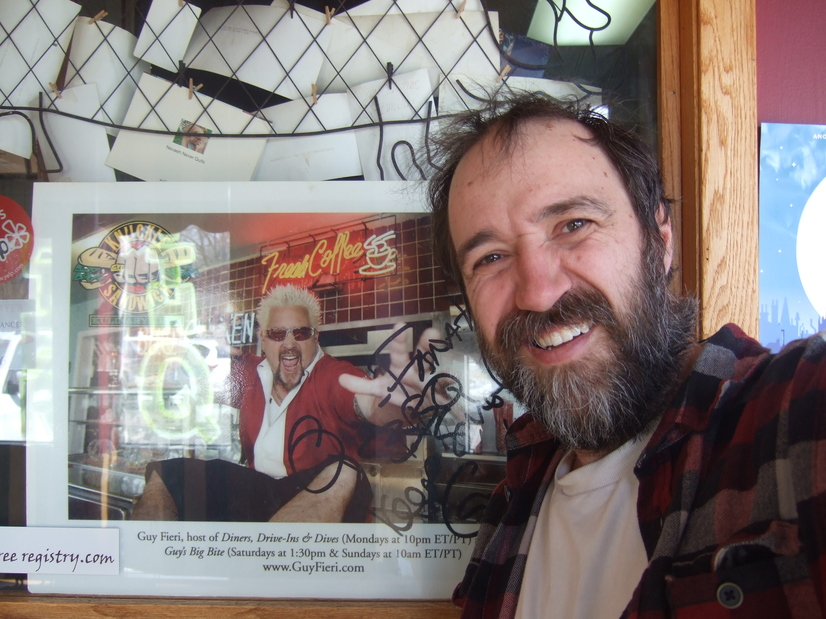 The author, a bearded man in a plaid shirt, smiling at the camera, standing next to a photo of Guy Fieri from Diners, Drive-Ins, and Dives.